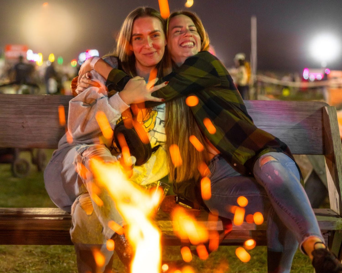 Two women hugging on a wooden bench at night by a campfire, with bright orange flames and glowing embers in the foreground. One woman wears a dark green plaid flannel shirt and light blue jeans, and the other wears a light gray hoodie and beige pants.