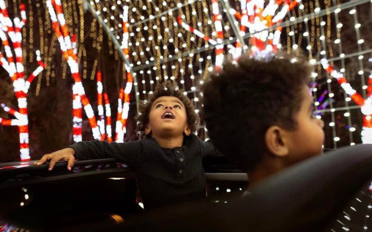 A young boy in a dark long-sleeve shirt looks up with a wide-eyed expression inside a dark ride vehicle, with another child in the foreground and festive red, white, and gold string lights hanging in the background.