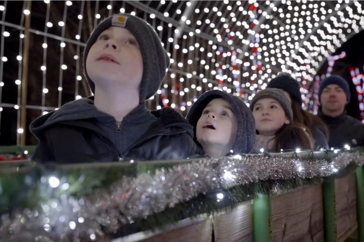A family wearing winter hats and jackets looks up at a canopy of glowing Christmas string lights in a festive nighttime setting.
