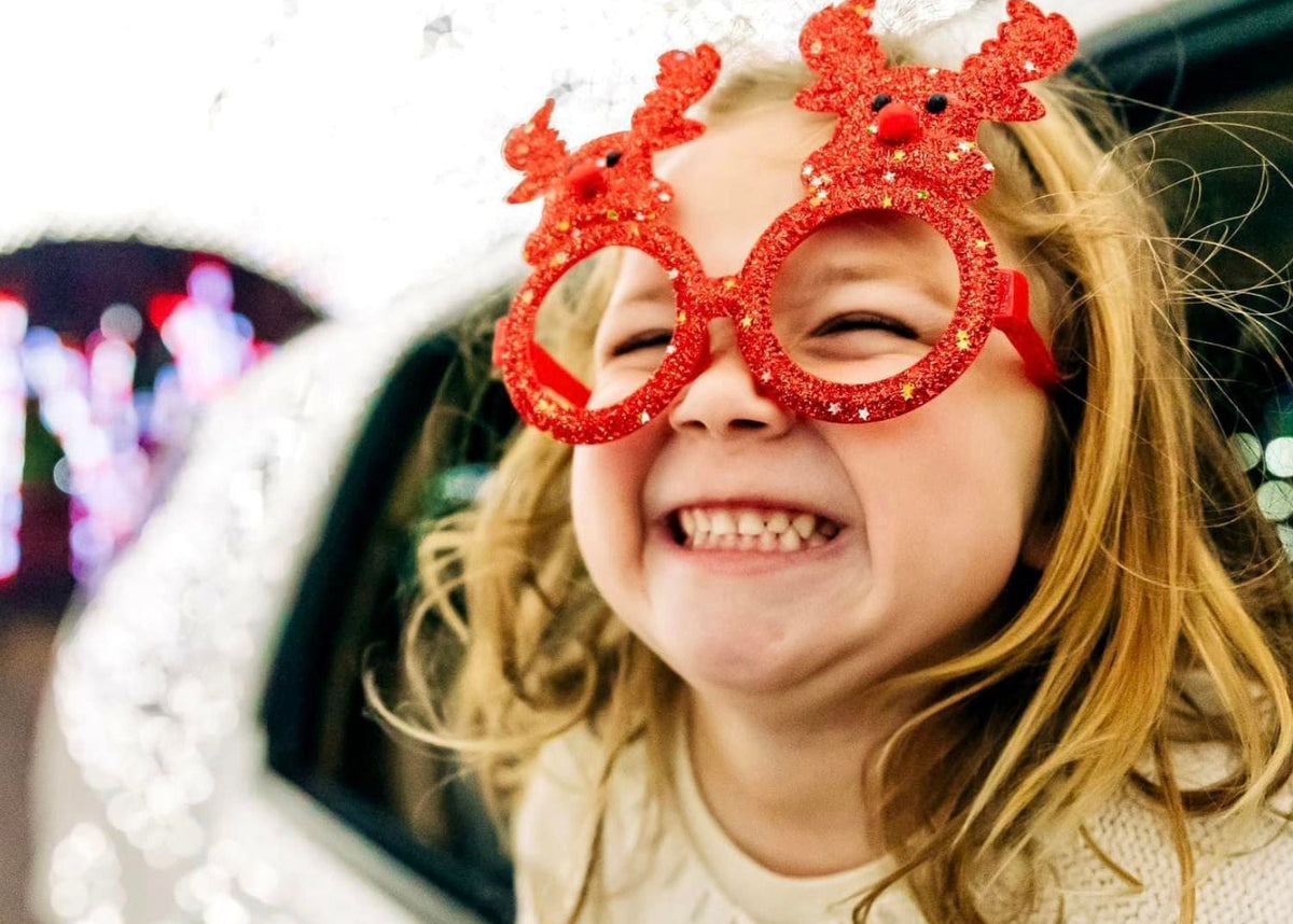 A smiling young girl with blonde hair wears red glittery round novelty glasses shaped like reindeer antlers, photographed outdoors near a white car with festive lights in the background.
