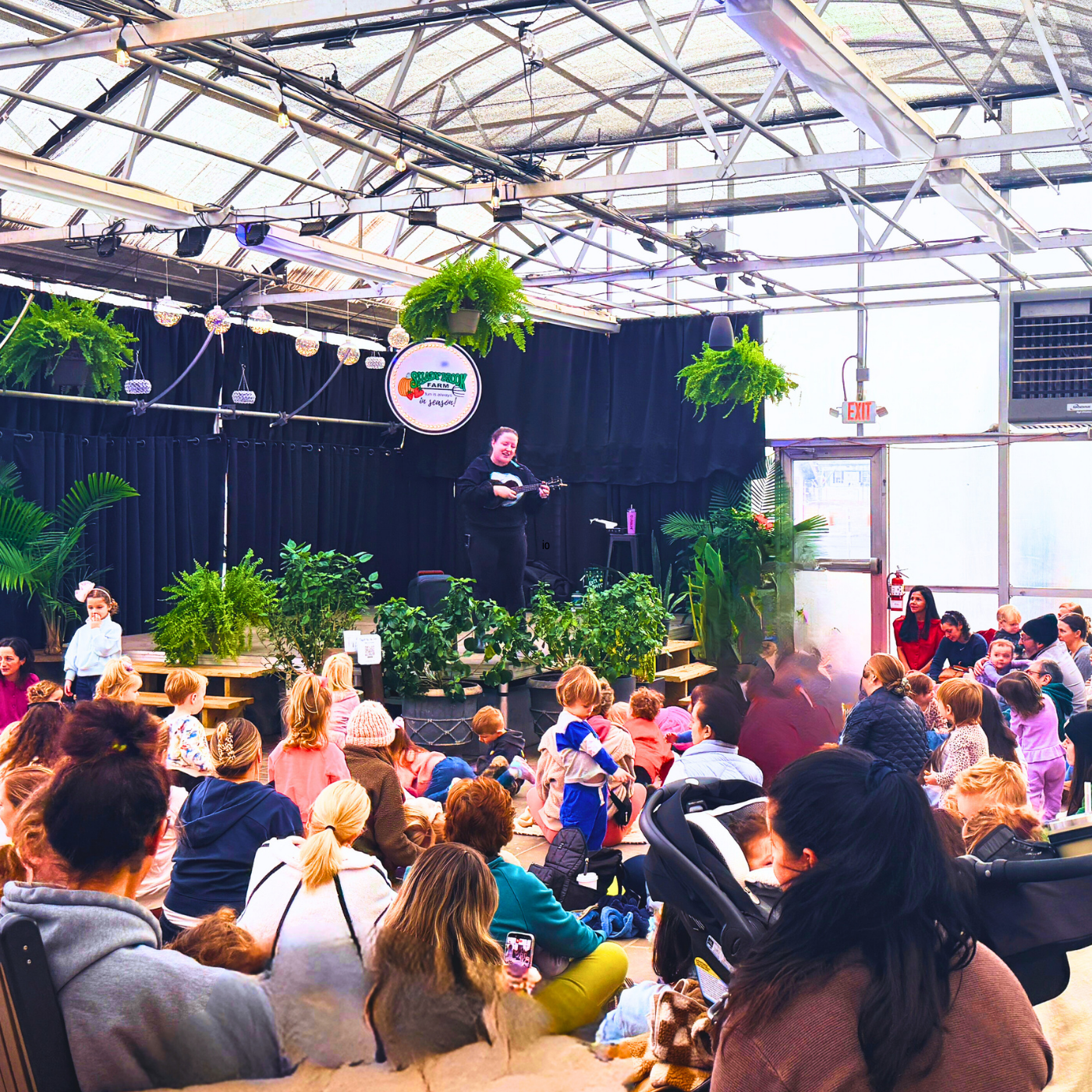 Person on stage with a crowd of people in an indoor setting with plants and a large screen.