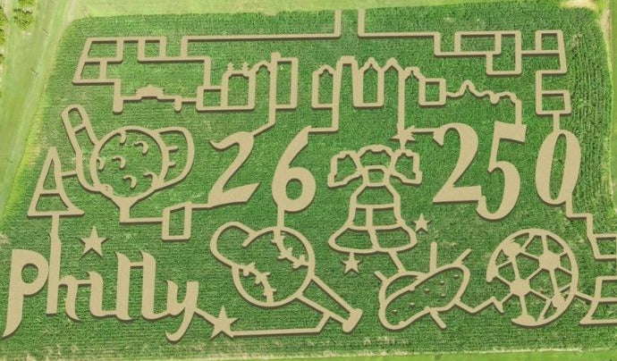 An aerial view of a large corn maze artwork in a green field, featuring the word 'Philly', the numbers 26 and 250, and various cartoon-style icons including a donut, angel, bell, and stars.