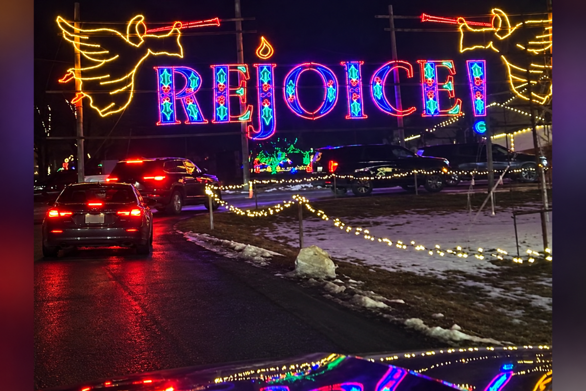 A nighttime street scene with a large illuminated decorative sign reading "REJOICE" in multicolored lights, flanked by angel figures. Cars with brake lights are visible on a wet road, and snow patches and string lights appear along the roadside.