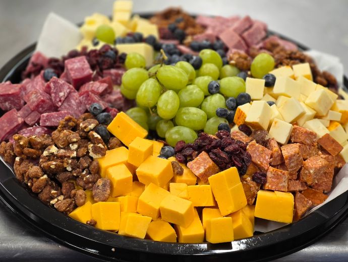 A charcuterie board platter filled with sliced salami, cheddar cheese cubes, dried cranberries, green grapes, blueberries, walnuts, and sliced meats on a black tray.