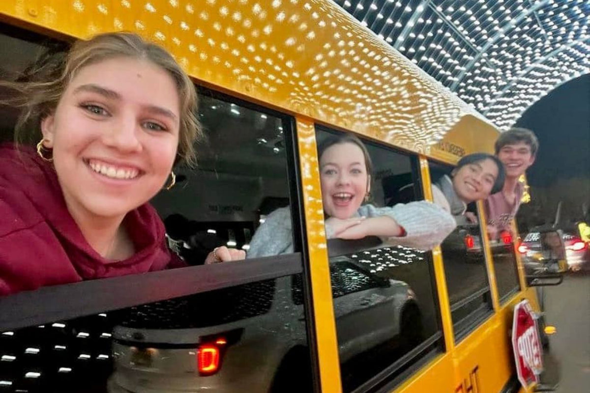 A yellow school bus at night with several young people leaning out of the windows. The bus is decorated with white string lights, and other vehicles and lights are visible in the background.