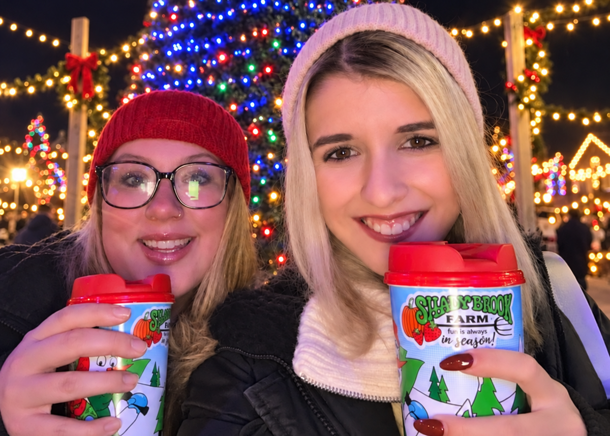 Two women smiling at night in front of a decorated Christmas tree, each holding a reusable coffee cup with a red lid and a holiday farm illustration. One wears a red beanie and glasses, and the other wears a light pink beanie and black jacket.