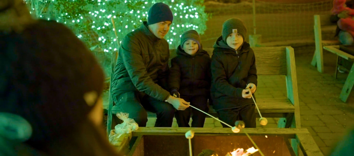 Three people in dark winter jackets and beanies sit around a campfire at night, roasting marshmallows on long handles. The scene is lit by warm firelight and green holiday string lights in the background.