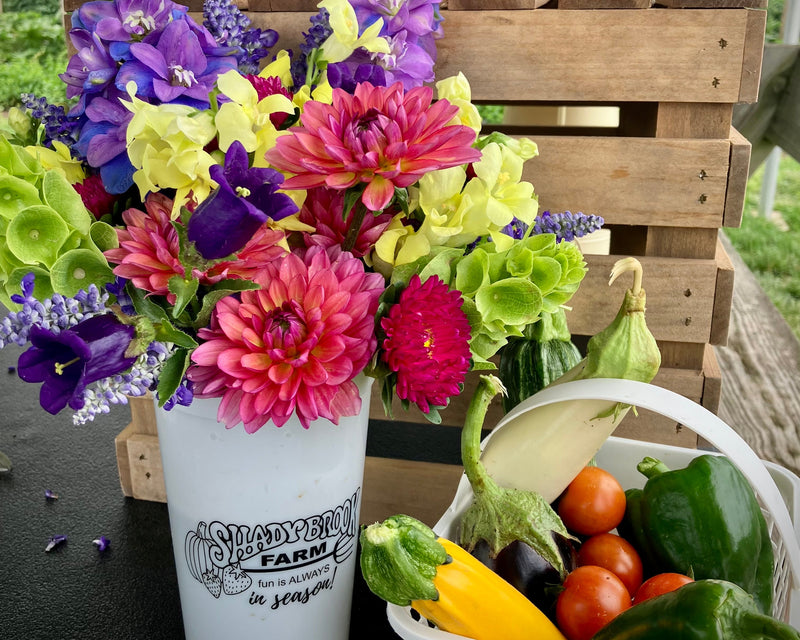 Colorful bouquet of flowers in a white container with a basket of vegetables on a wooden crate.
